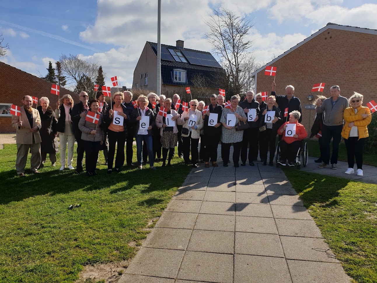 Medlemmer af Selmersbo står samlet udenfor med danske flag og bogstavskilte, der staver Selmersbo